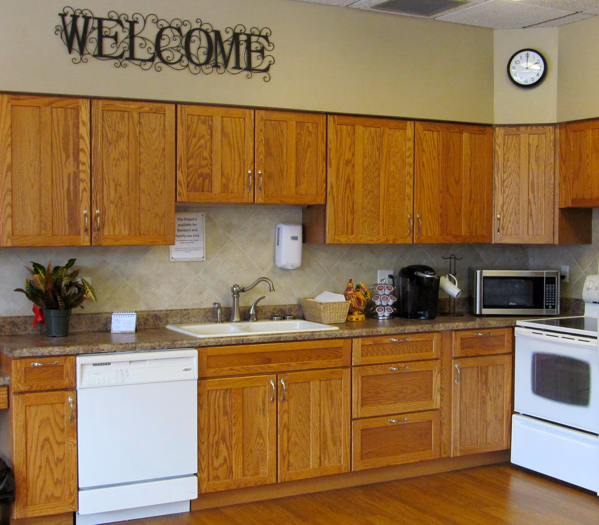A kitchen area with wooden cabinets, a white dishwasher, a sink with a faucet, a coffee maker, a microwave, and a white stove. There is a plant on the counter, a clock on the wall showing 12:10, and a decorative metal sign that says 'WELCOME' above the cabinets.