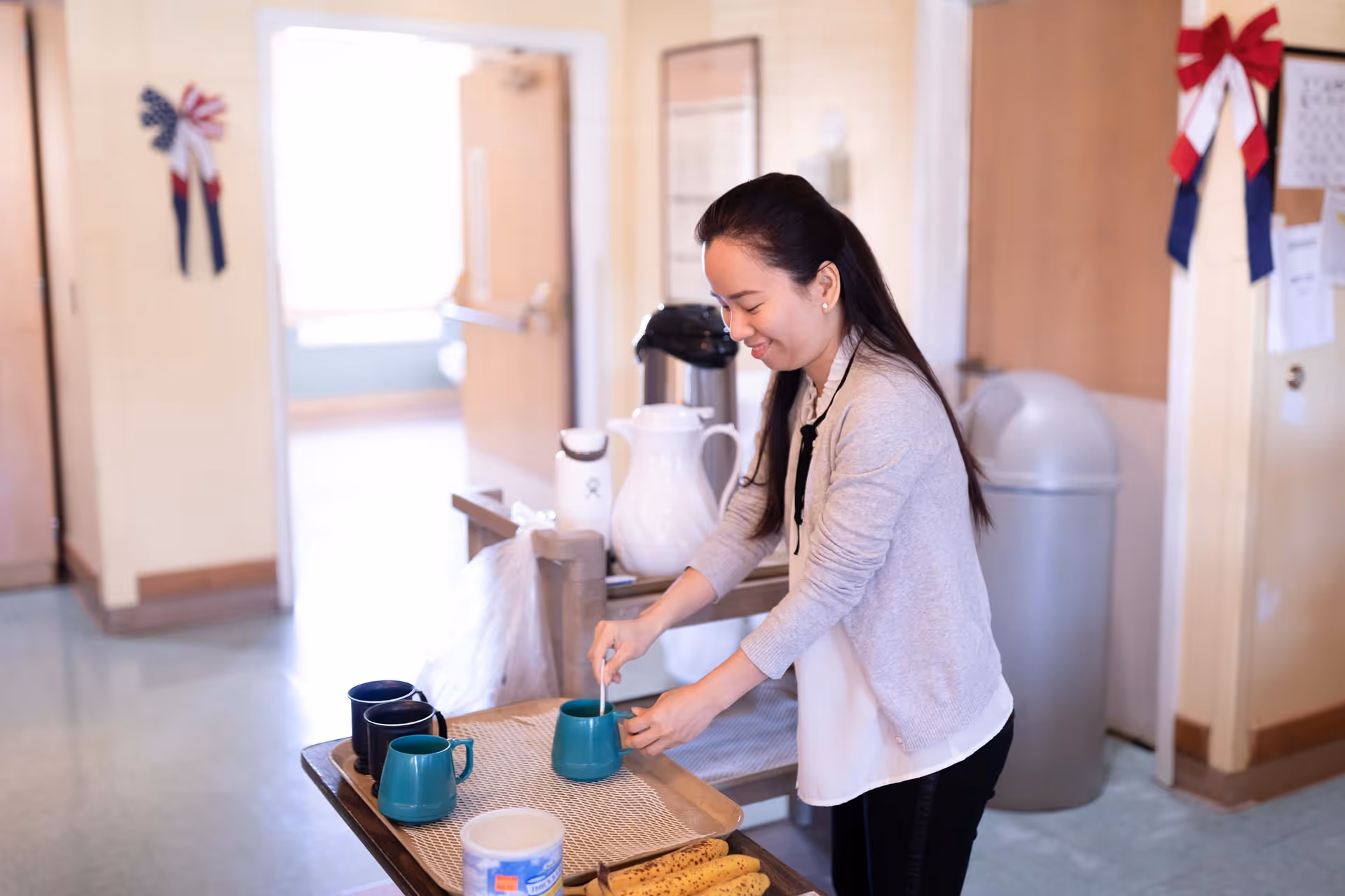 A woman in a light gray cardigan and white blouse is preparing coffee or tea at a serving cart in a nursing and rehabilitation center. The cart has several mugs, a white pitcher, a coffee dispenser, and some snacks. The background shows a hallway with open doors and some wall decorations including red, white, and blue ribbons.