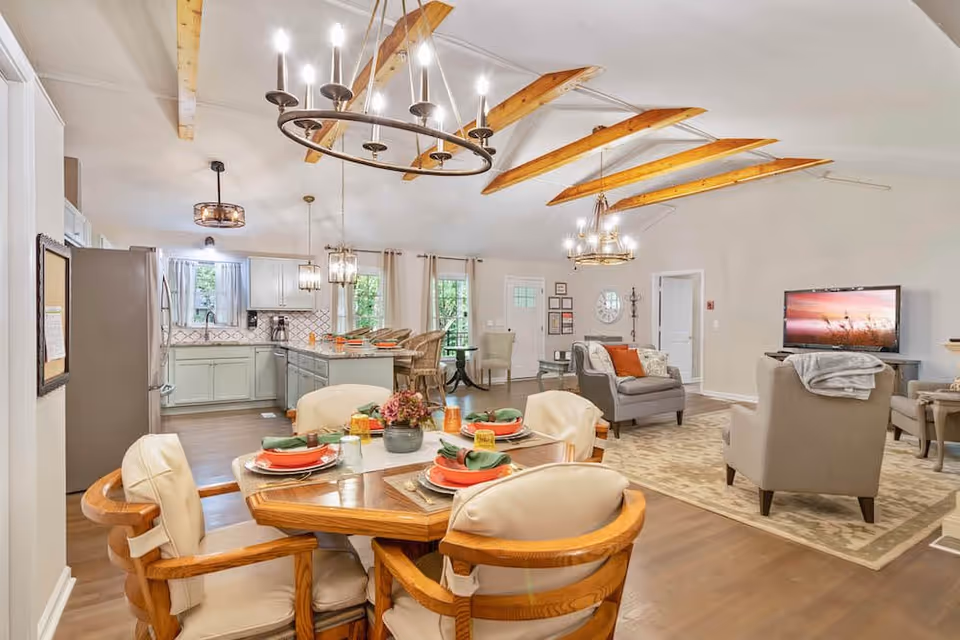 A spacious and well-lit senior living common area featuring a dining table set for four with plates, napkins, and a centerpiece. The room includes a kitchen with light-colored cabinets, a refrigerator, and a countertop with bar stools. The living area has comfortable armchairs, a rug, and a TV mounted on a stand. The ceiling has exposed wooden beams and multiple light fixtures, and large windows with curtains allow natural light to enter.