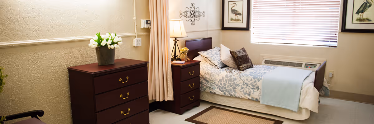 Single-occupancy bedroom with a neatly made bed, bedside table and lamp, dresser topped with flowers, window with blinds, and neutral decor.