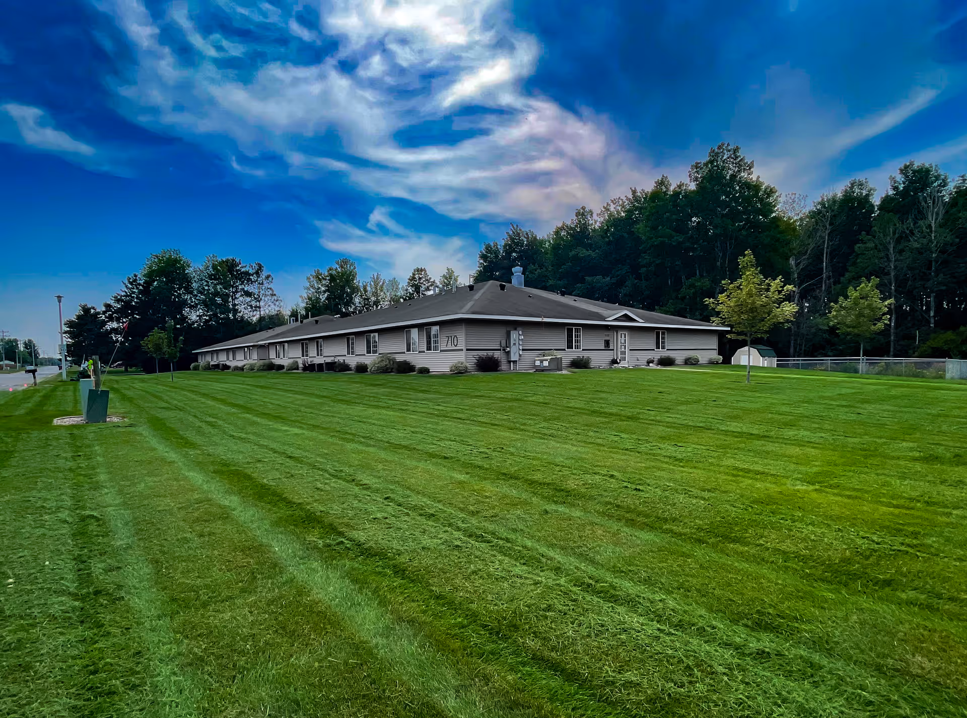 Long single-story residential building with a wide mowed lawn and trees under a dramatic cloudy sky.