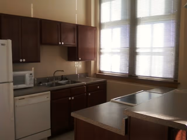 A small apartment kitchen with dark wood cabinets, a double sink, dishwasher, microwave, refrigerator, and a countertop island by windows with blinds.