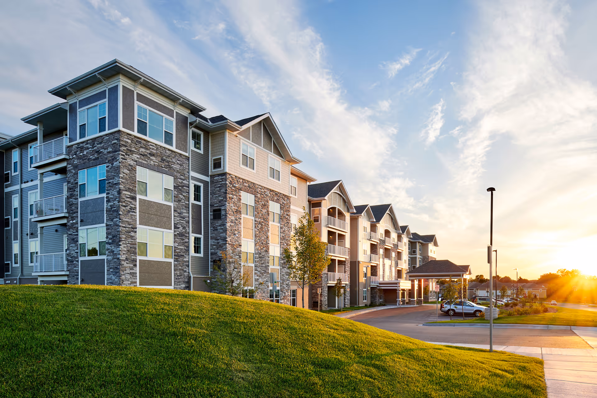 Exterior view of a modern multi-story senior living facility building with stone and siding facade, balconies, a driveway with parked cars, green lawn in the foreground, and a sunset sky in the background.