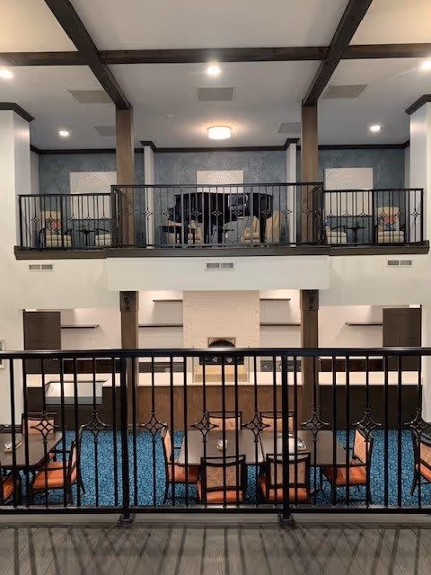 Interior view of a two-level common area in a senior living facility. The lower level features a seating area with tables and chairs on a blue carpet, and a fireplace centered on the far wall. The upper level has a balcony with black metal railings, seating arrangements including chairs and a piano, and decorative wall panels. The ceiling has exposed wooden beams and recessed lighting.
