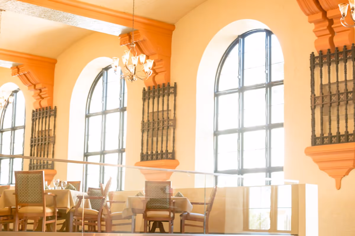 Sunlit dining area with arched windows, chandeliers, and set tables and chairs.