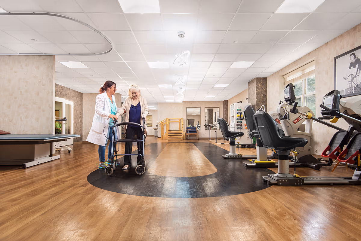 A rehabilitation room with exercise equipment and a wooden floor. A healthcare professional is assisting an elderly woman using a walker. The room is well-lit with ceiling lights and has a large window on the right side.