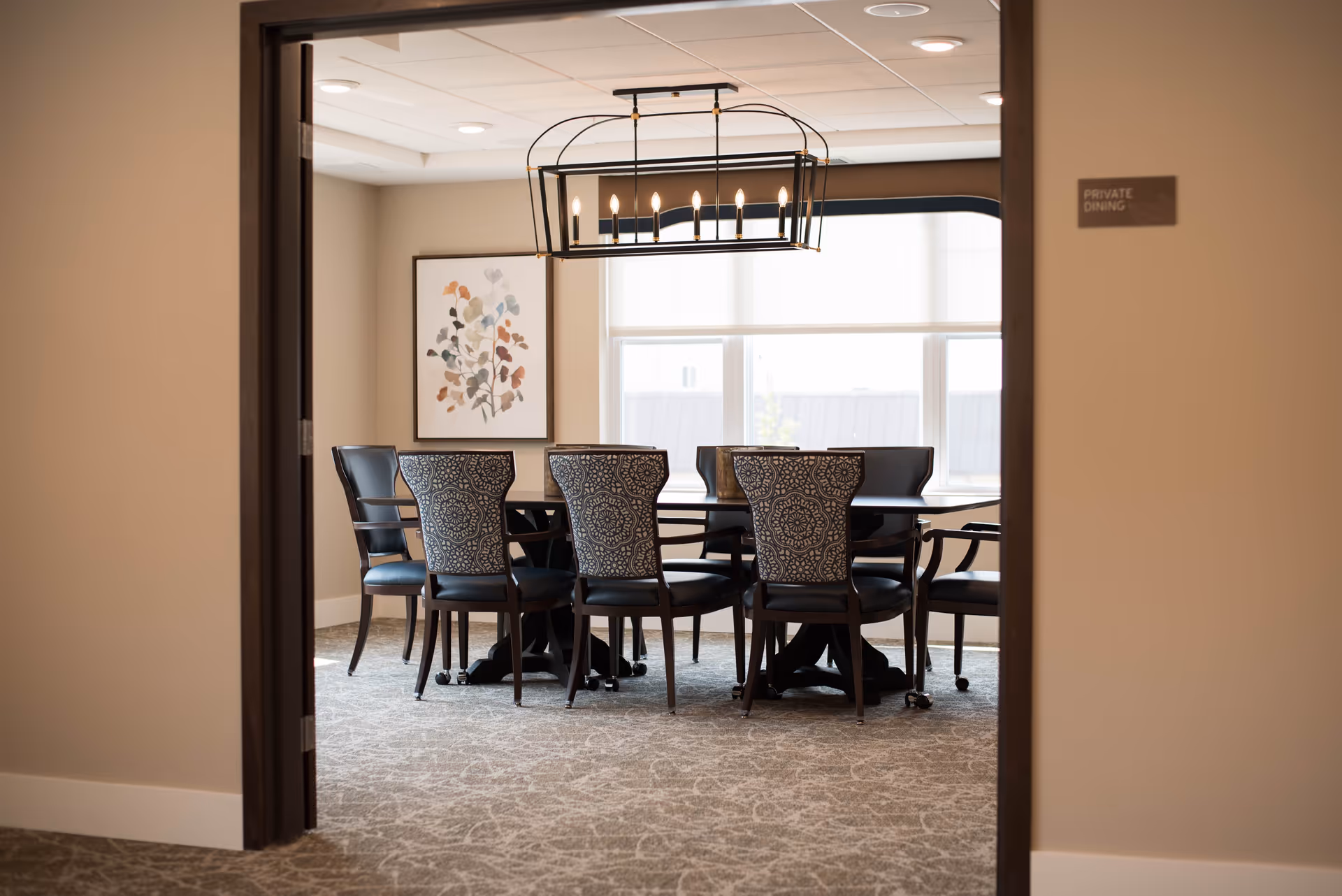 View through an open doorway into a private dining room with a rectangular table surrounded by eight chairs. The chairs have patterned backs and black seats. A modern chandelier with exposed bulbs hangs above the table. A large window with white blinds lets in natural light, and a framed artwork with abstract floral design is on the wall.