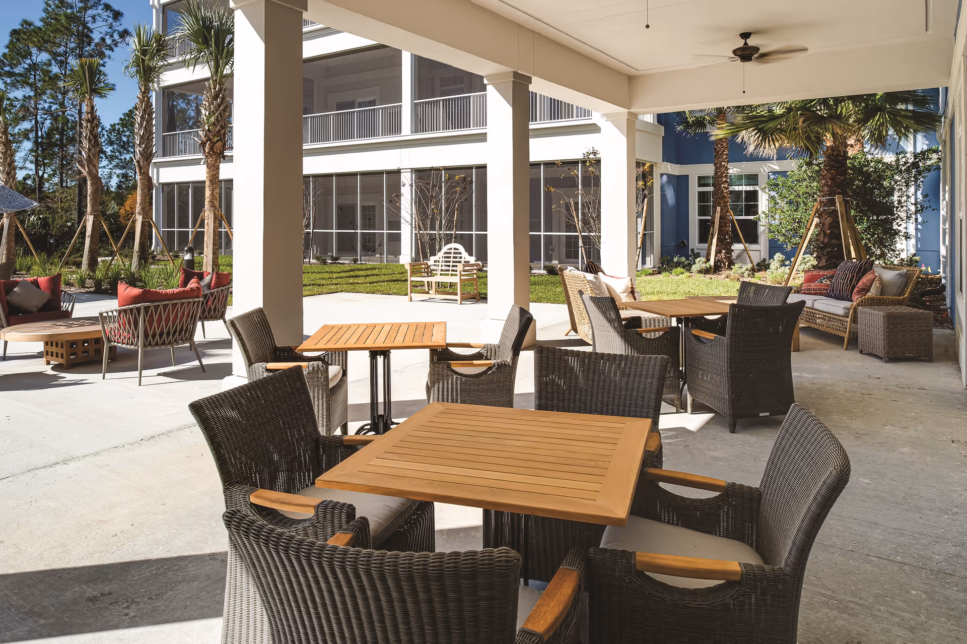 Covered outdoor patio area with multiple wooden tables and wicker chairs with cushions. In the background, there are palm trees, green grass, and a building with large windows and balconies.