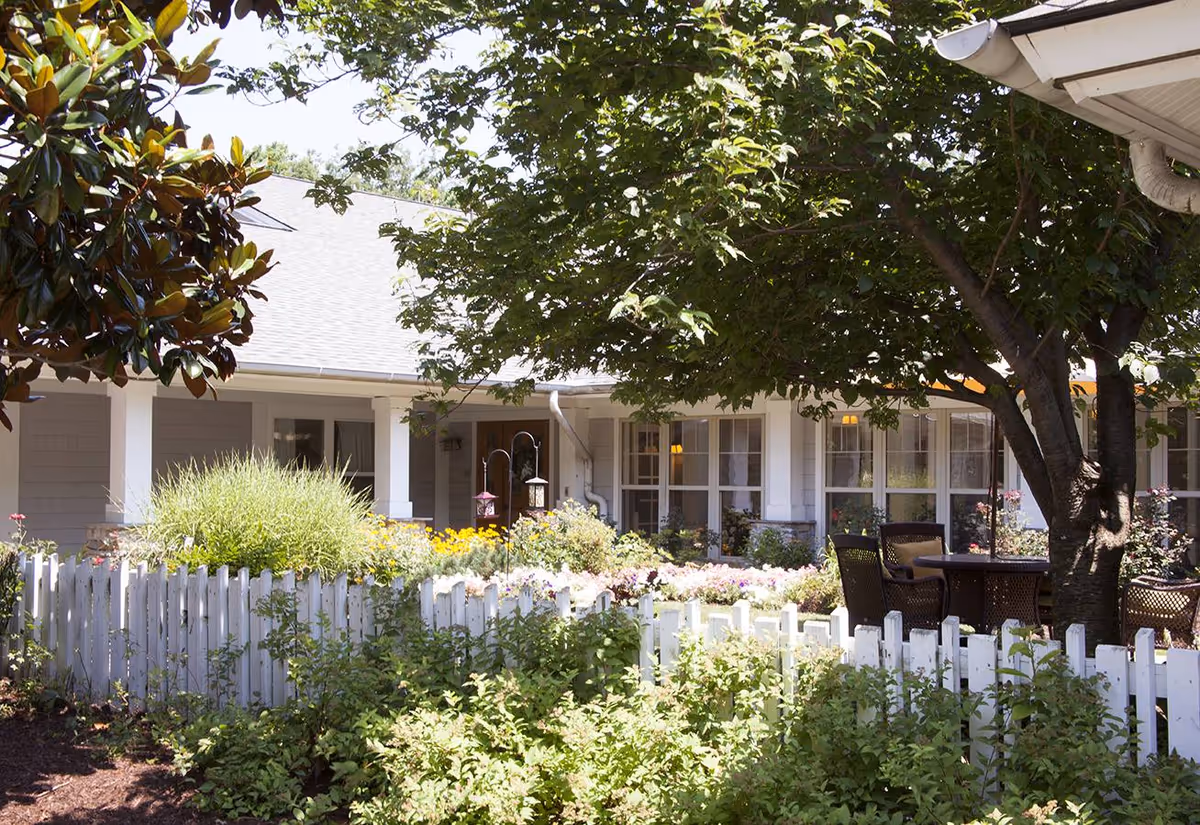 Outdoor garden area at a senior living facility with a white picket fence, lush green plants, colorful flowers, and a large tree providing shade over a patio table with chairs. The building with multiple windows and a covered porch is visible in the background.
