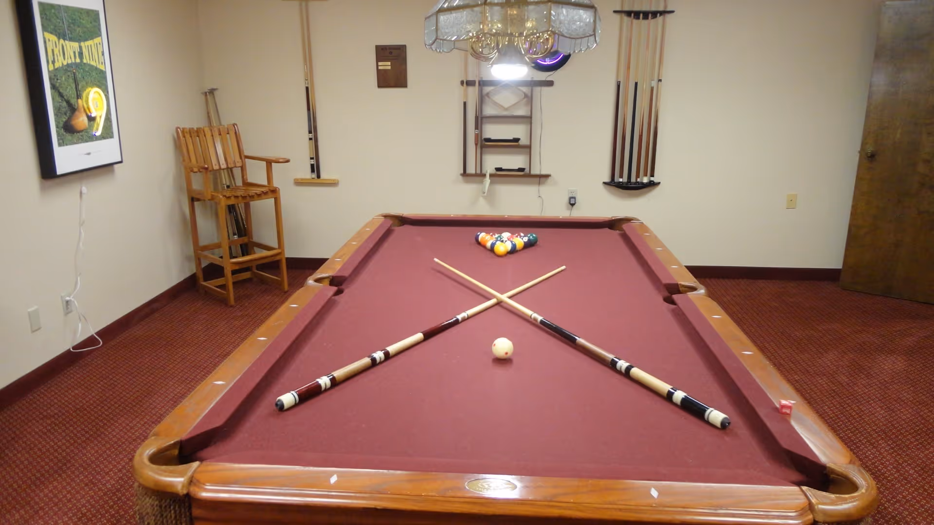 Indoor recreation room featuring a burgundy pool table with two crossed cues and a racked set of balls, with seating and cue storage along the walls.