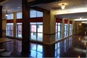 Interior hallway of a senior living facility with large windows along the walls, polished floors, and ceiling lights. The hallway has a warm, inviting atmosphere with red curtains on the windows and a long corridor extending into the distance.