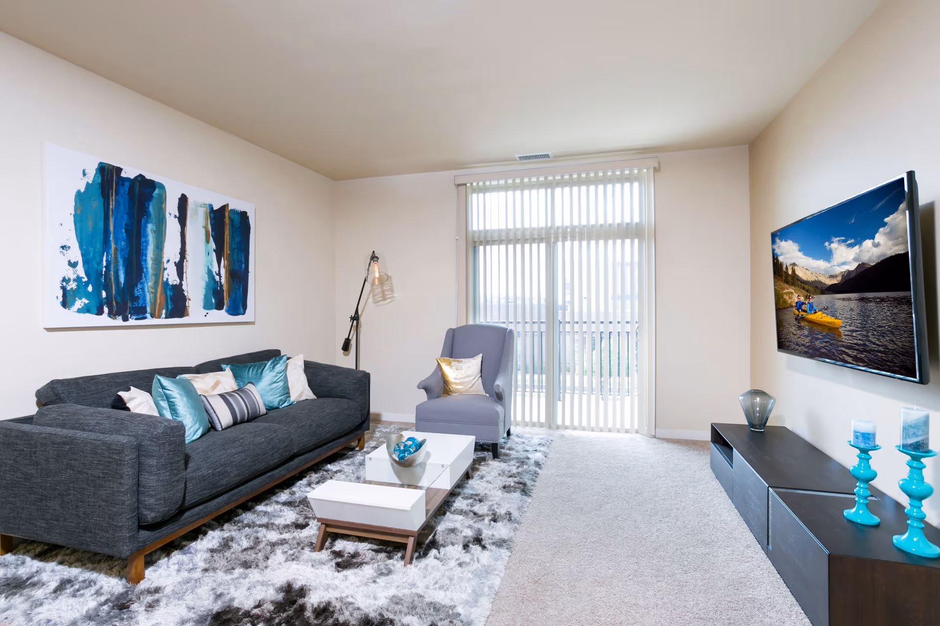 A modern living room with a dark gray sofa adorned with teal and white pillows, a gray armchair with a gold pillow, a white coffee table on a fluffy gray and white rug, a floor lamp, and a wall-mounted TV displaying a scenic image of people kayaking on a lake surrounded by mountains. Vertical blinds cover the large window behind the armchair.