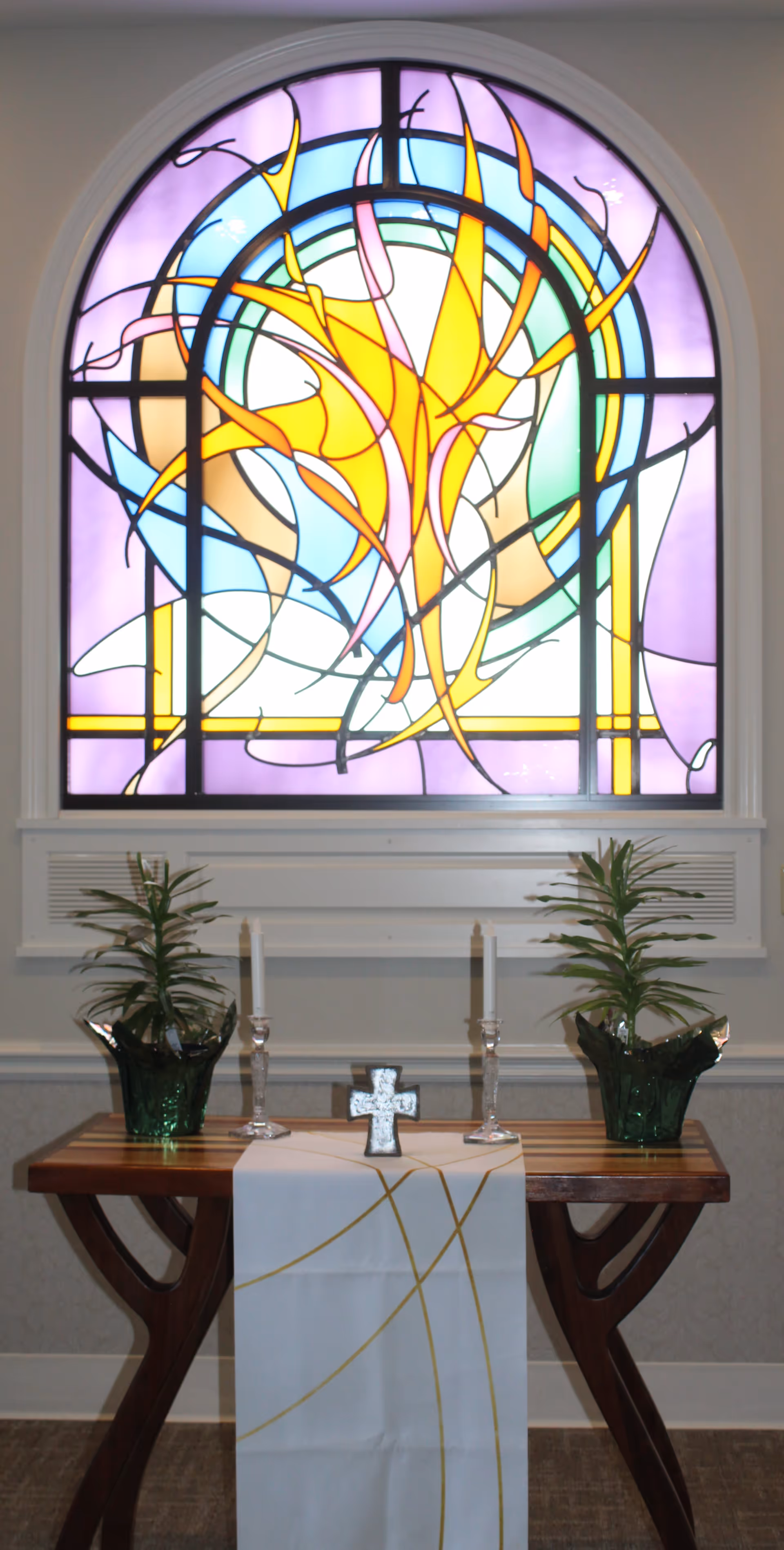 A wooden table with a white cloth featuring gold lines draped over it, holding a small silver cross and two white candles in glass holders. On either side of the table are green potted plants. Behind the table is a large arched stained glass window with abstract colorful designs in yellow, blue, purple, and green.