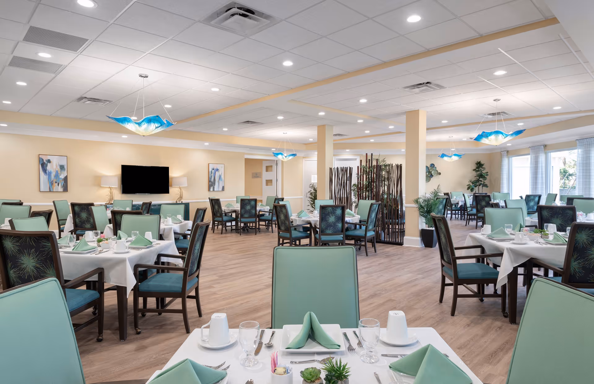 A spacious dining room with multiple tables covered in white tablecloths, each set with green napkins, cups, glasses, and silverware. The chairs have green upholstery with dark wooden frames. The room features light-colored walls, wood flooring, modern blue pendant lights, and large windows letting in natural light. There are decorative plants and artwork on the walls, and a flat-screen TV mounted on one wall.