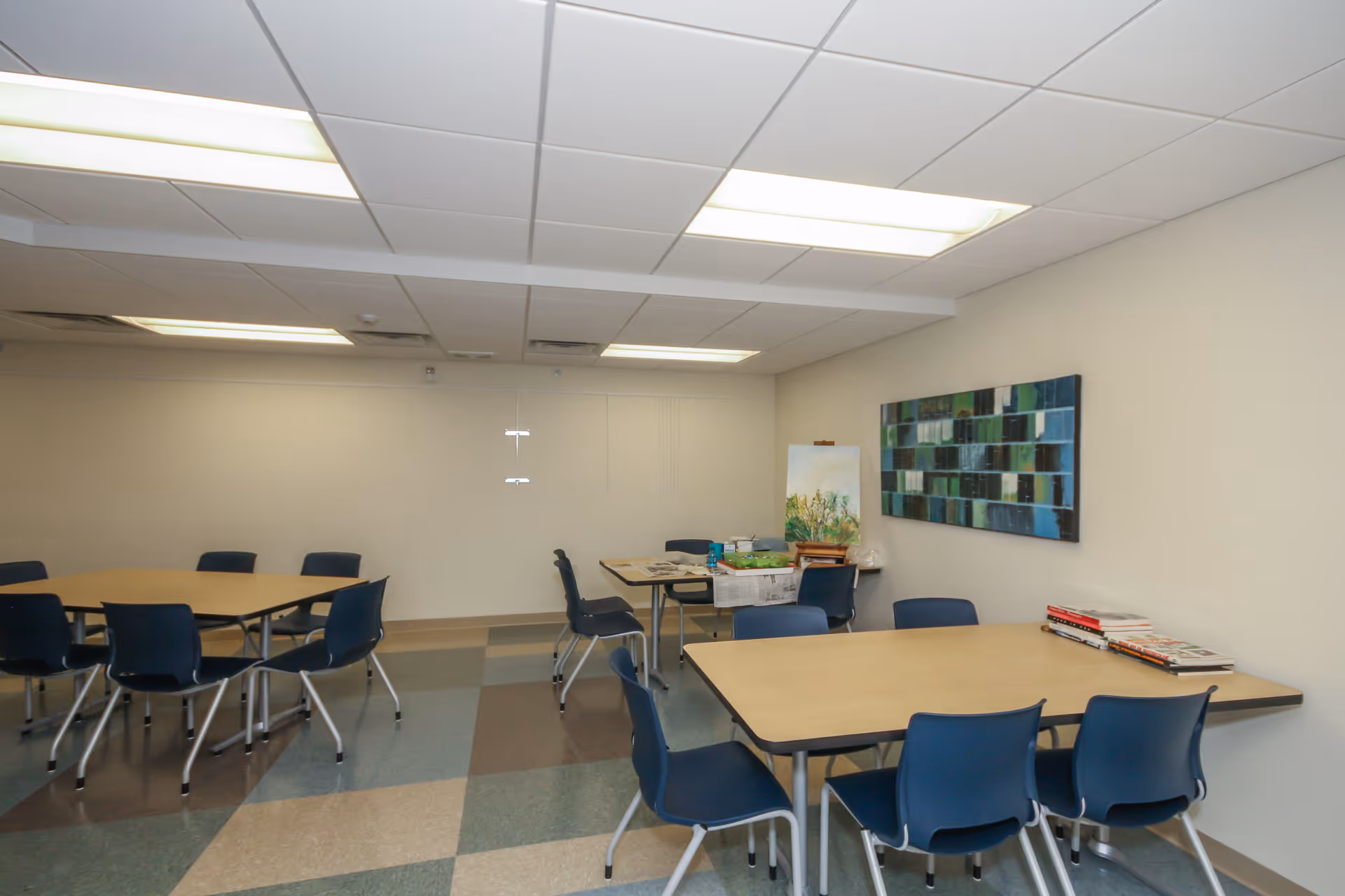 A room with several tables and blue chairs arranged around them. There is a colorful abstract painting on the wall and some books and art supplies on one of the tables. The floor has a checkered pattern with different shades of blue and beige tiles. The ceiling has fluorescent lights.