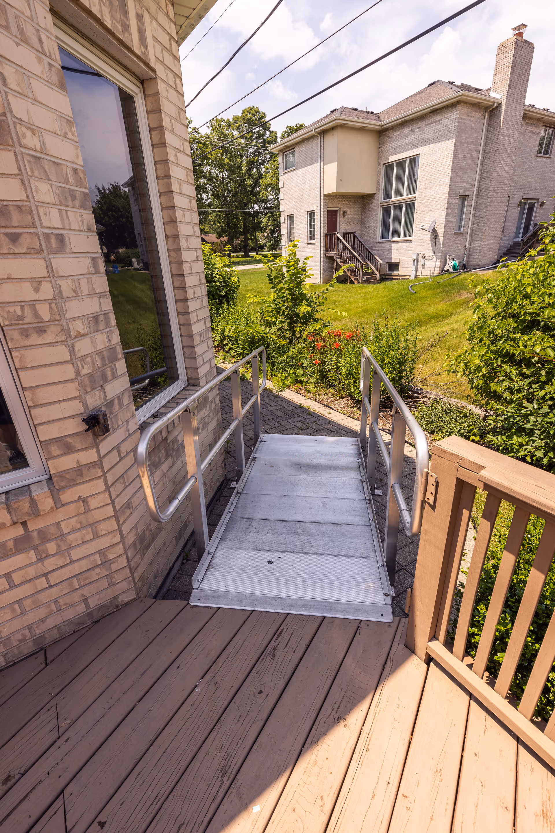 Outdoor view of a metal wheelchair ramp attached to a wooden deck leading to a brick building entrance, with a grassy yard, bushes, and another house visible in the background under a partly cloudy sky.