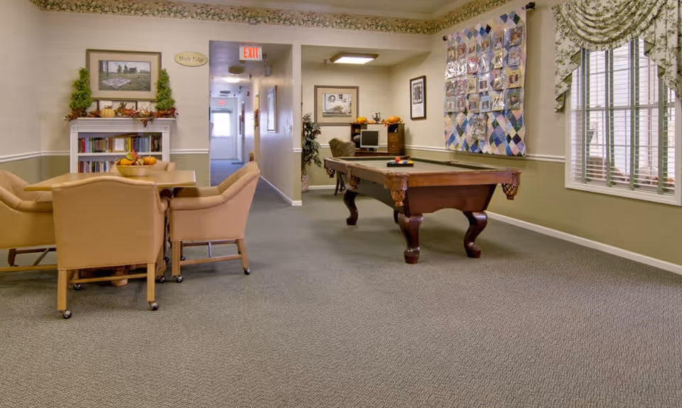 A senior living common room with a pool table, a table and chairs, a bookshelf, computer desk and window treatments.