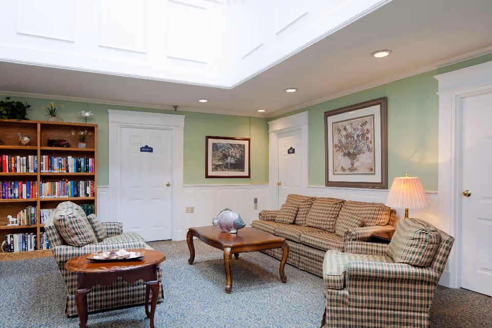 A cozy living room area with a plaid sofa and two matching armchairs around a wooden coffee table. There is a wooden bookshelf filled with books on the left side, two framed pictures on the green walls, and a table lamp on a side table next to the sofa. The room has a skylight ceiling and three white doors with signs on them.