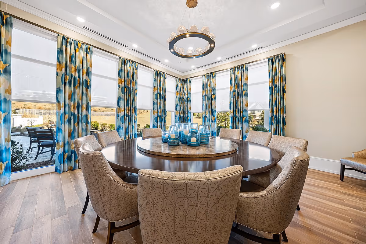 A bright dining room with a large round wooden table surrounded by beige patterned chairs. The table has a circular marble centerpiece with blue glass candle holders. The room features tall windows with white blinds and blue, yellow, and white patterned curtains. A modern circular chandelier hangs from the ceiling, and the floor is light wood.