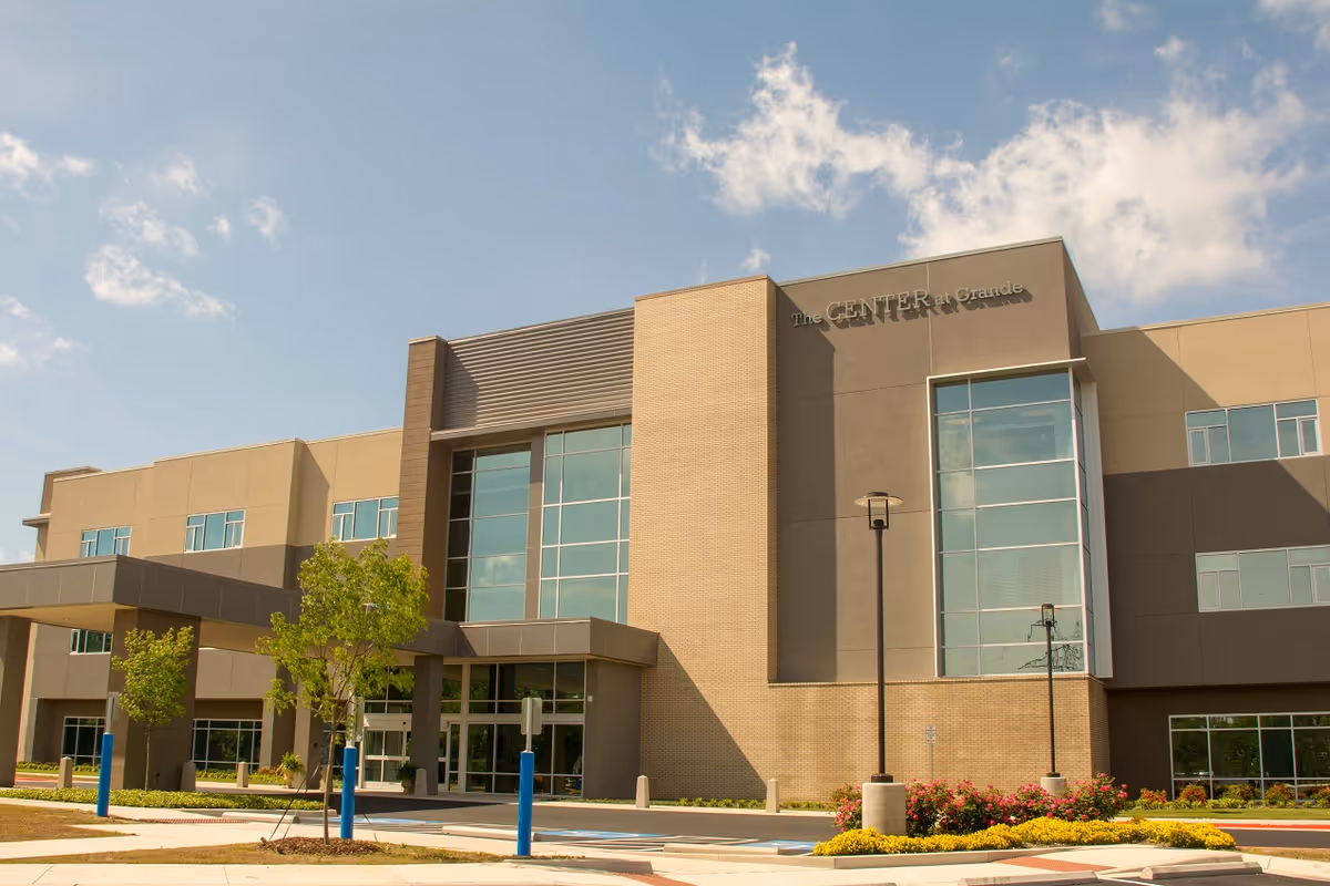 Exterior view of a modern three-story building named The Center At Grande with large windows, a covered entrance, landscaped greenery, and a clear blue sky with some clouds.