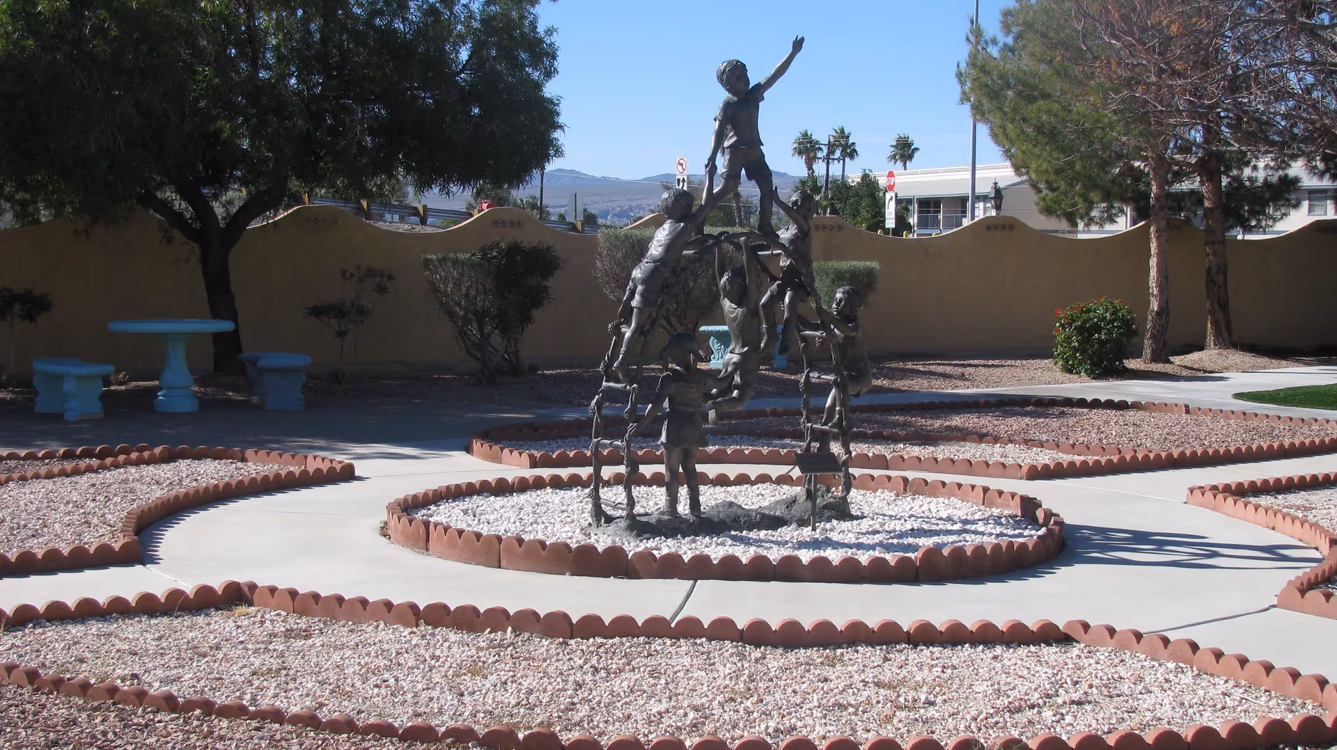 Outdoor garden area with a bronze sculpture of children playing and climbing on each other, surrounded by circular and curved pathways bordered with red bricks and filled with white gravel. There are trees, bushes, and a beige stucco wall in the background, along with a blue table and benches under a tree.