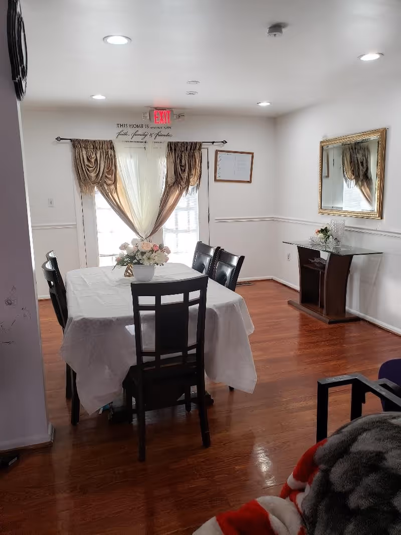 A dining room with a rectangular table covered with a white tablecloth and six dark wooden chairs around it. A vase with flowers is placed in the center of the table. The room has wooden flooring, white walls, and a large window with sheer and brown curtains. There is a decorative console table with a glass top and a large mirror with a gold frame on the right wall. An exit sign is visible above the window, and a framed whiteboard hangs on the wall to the right of the window.