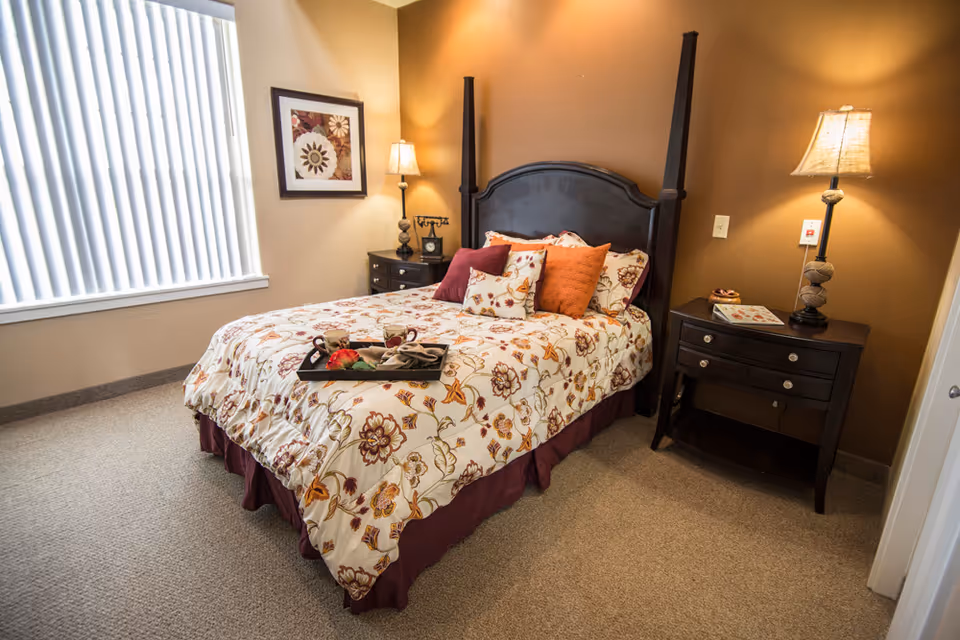Sunlit bedroom featuring a floral-covered bed with decorative pillows, two nightstands and lamps.