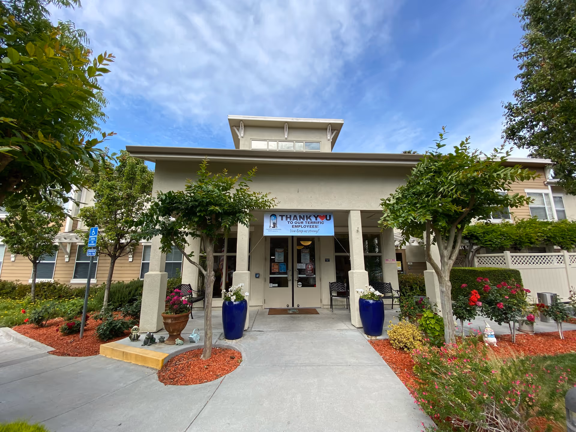 Front entrance of a senior living facility named Pacific Gardens with a covered walkway, two large blue planters with flowers, small trees, and landscaped garden beds with red mulch and various plants. A banner above the entrance thanks the employees. The sky is partly cloudy.