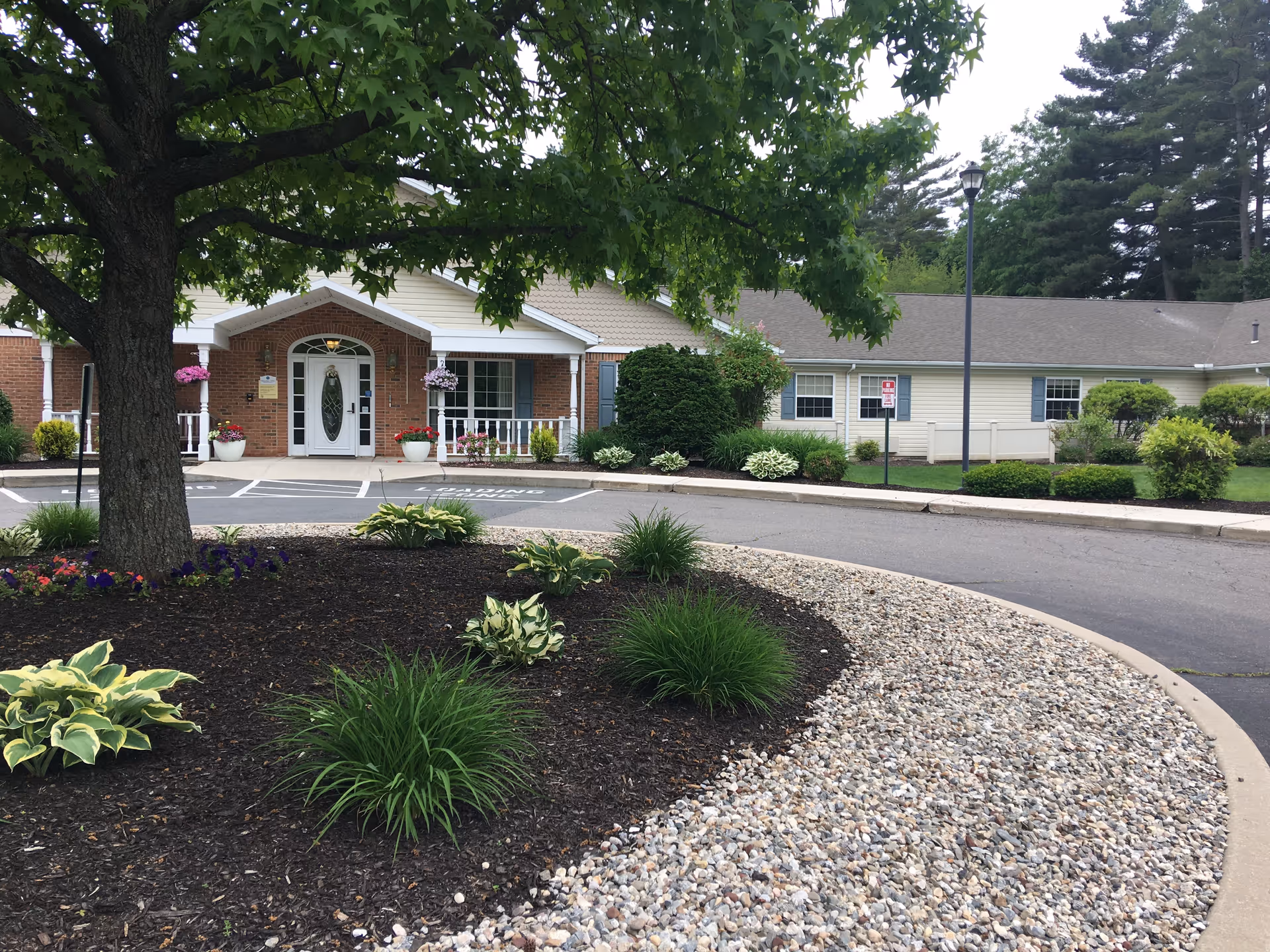 Front entrance of a single-story assisted living building with a circular driveway, landscaped beds, and hanging flower baskets.
