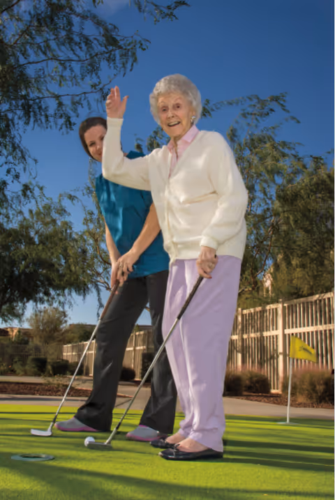 A senior woman is joyfully playing mini-golf with a caregiver in a bright sunny outdoor setting.