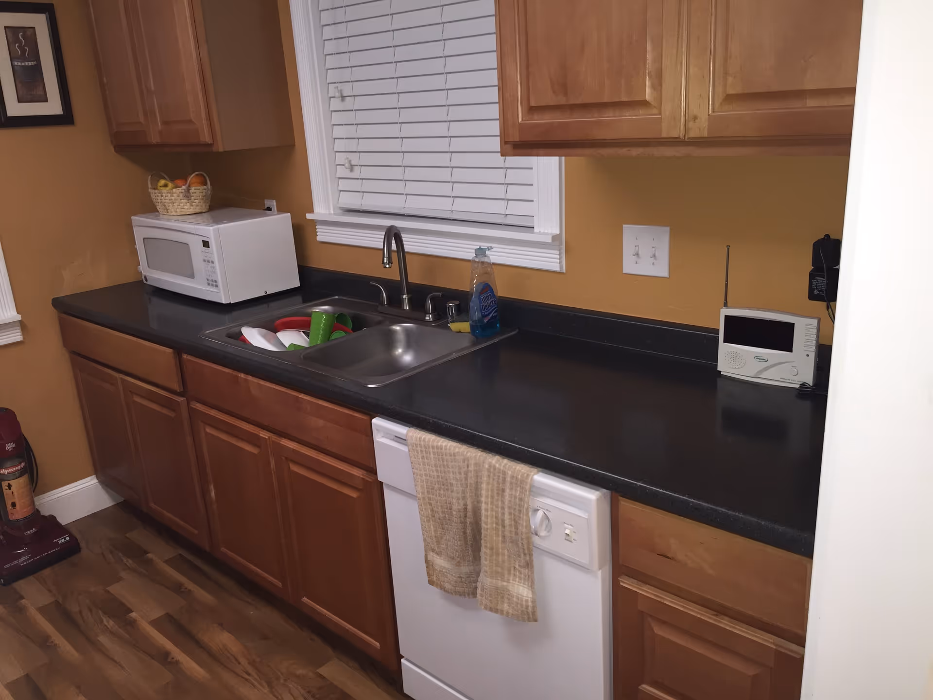 A kitchen area with wooden cabinets, a black countertop, a white microwave, a sink with dishes inside, a bottle of dish soap, a white dishwasher with a beige towel hanging on its handle, and a small electronic device on the counter. The walls are painted yellow, and there is a window with white blinds above the sink.