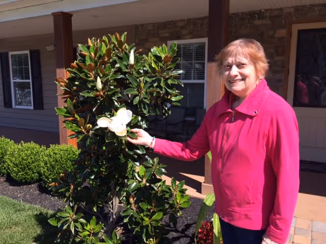 An elderly woman wearing a bright pink jacket stands outside near a flowering bush with large green leaves and white flowers, smiling at the camera. Behind her is a building with windows and a porch area.
