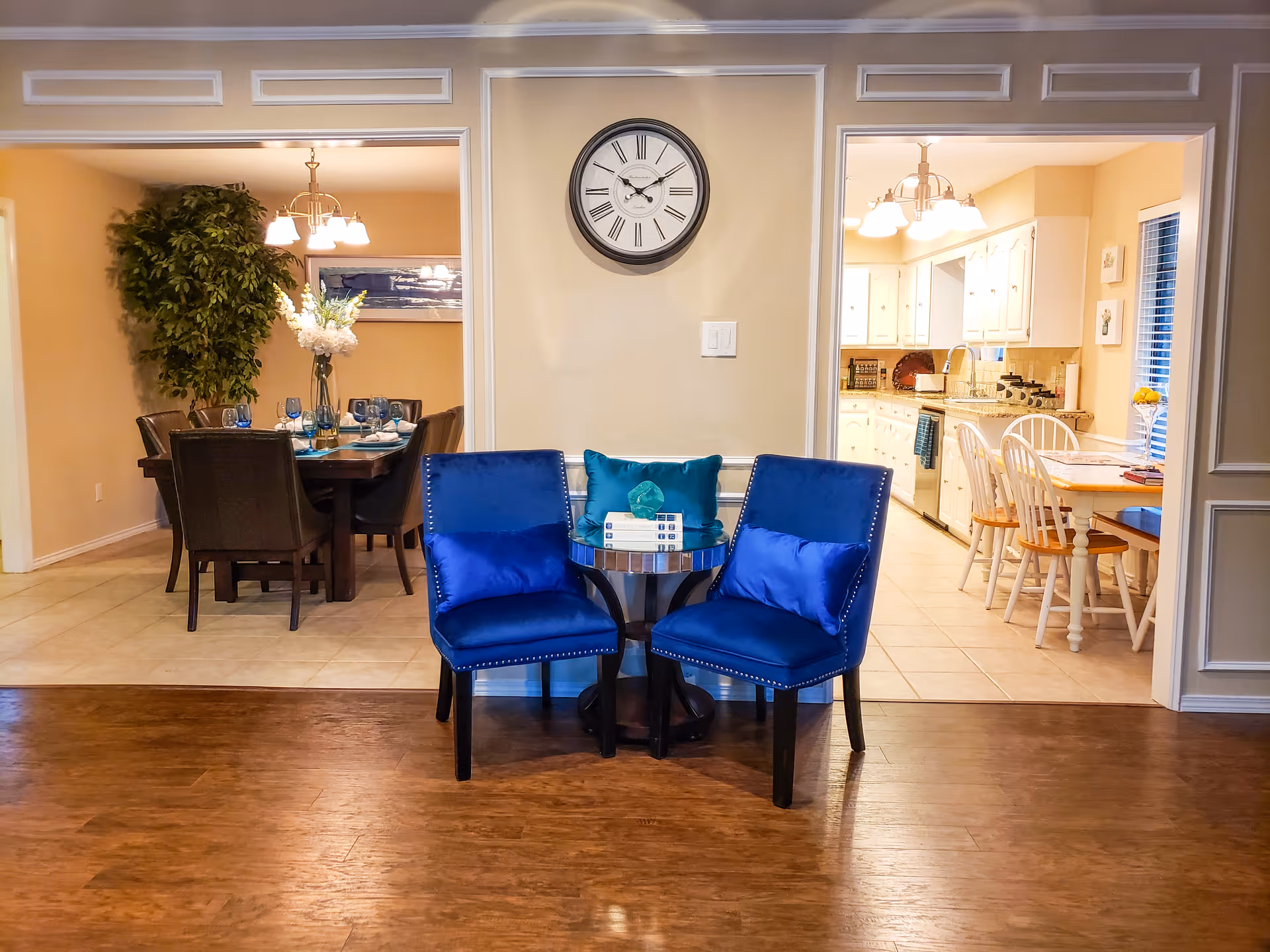 Two blue chairs and a small table beneath a wall clock in an interior common area with a dining room visible on the left and a kitchen on the right.