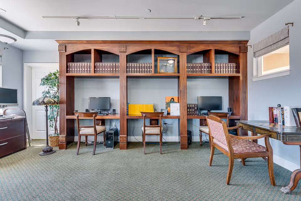 Interior common room with built-in wooden desks and bookshelves, computers and chairs arranged for residents.