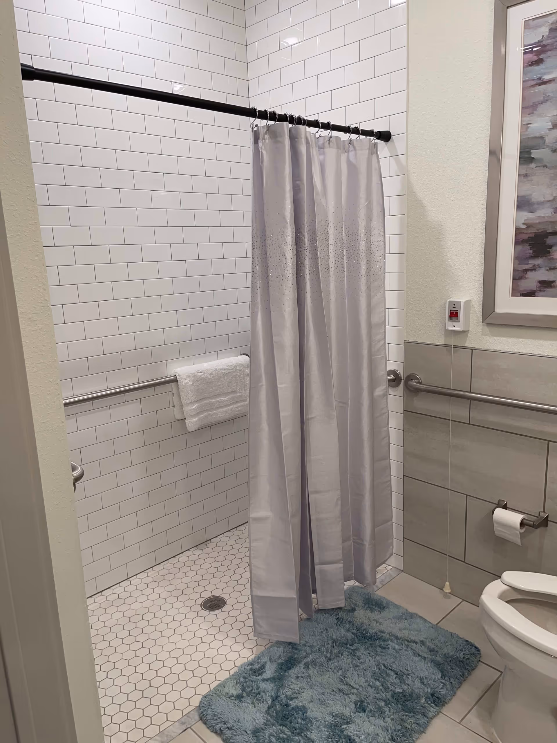 A bathroom with a walk-in shower featuring white subway tiles and a white shower curtain on a black rod. There is a white towel hanging on a metal grab bar inside the shower. The floor has small hexagonal white tiles. Outside the shower is a blue textured bath mat. On the right side, there is a toilet with a toilet paper holder mounted on the wall, and a framed abstract painting above the grab bar. A pull cord emergency alert device is mounted on the wall near the painting.