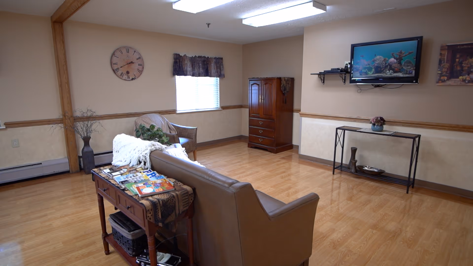 A seating area with a leather sofa, armchair, wall-mounted TV and small tables on polished wood floors in a common room.