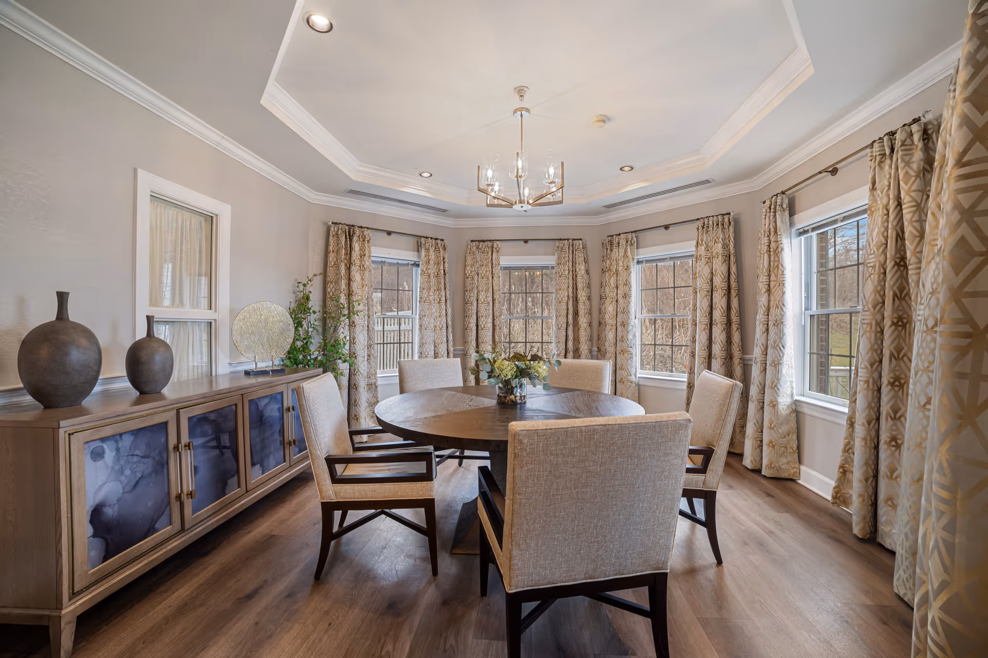 Elegant dining room with a round wooden table surrounded by upholstered chairs, a sideboard with decorative vases, chandelier, and multiple windows with patterned curtains.