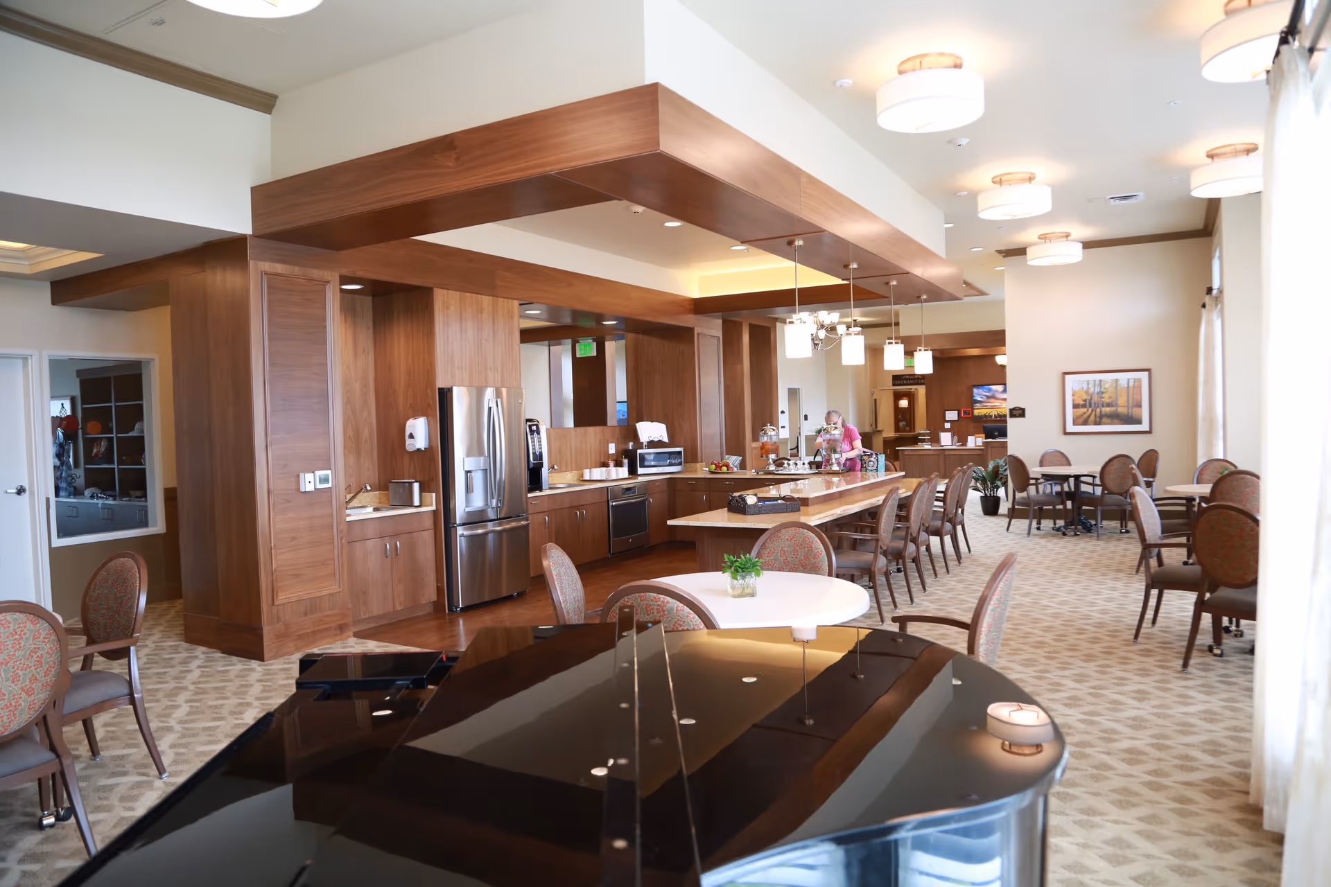 Bright communal dining and kitchenette area with a grand piano, round tables, a long island and wooden cabinetry.