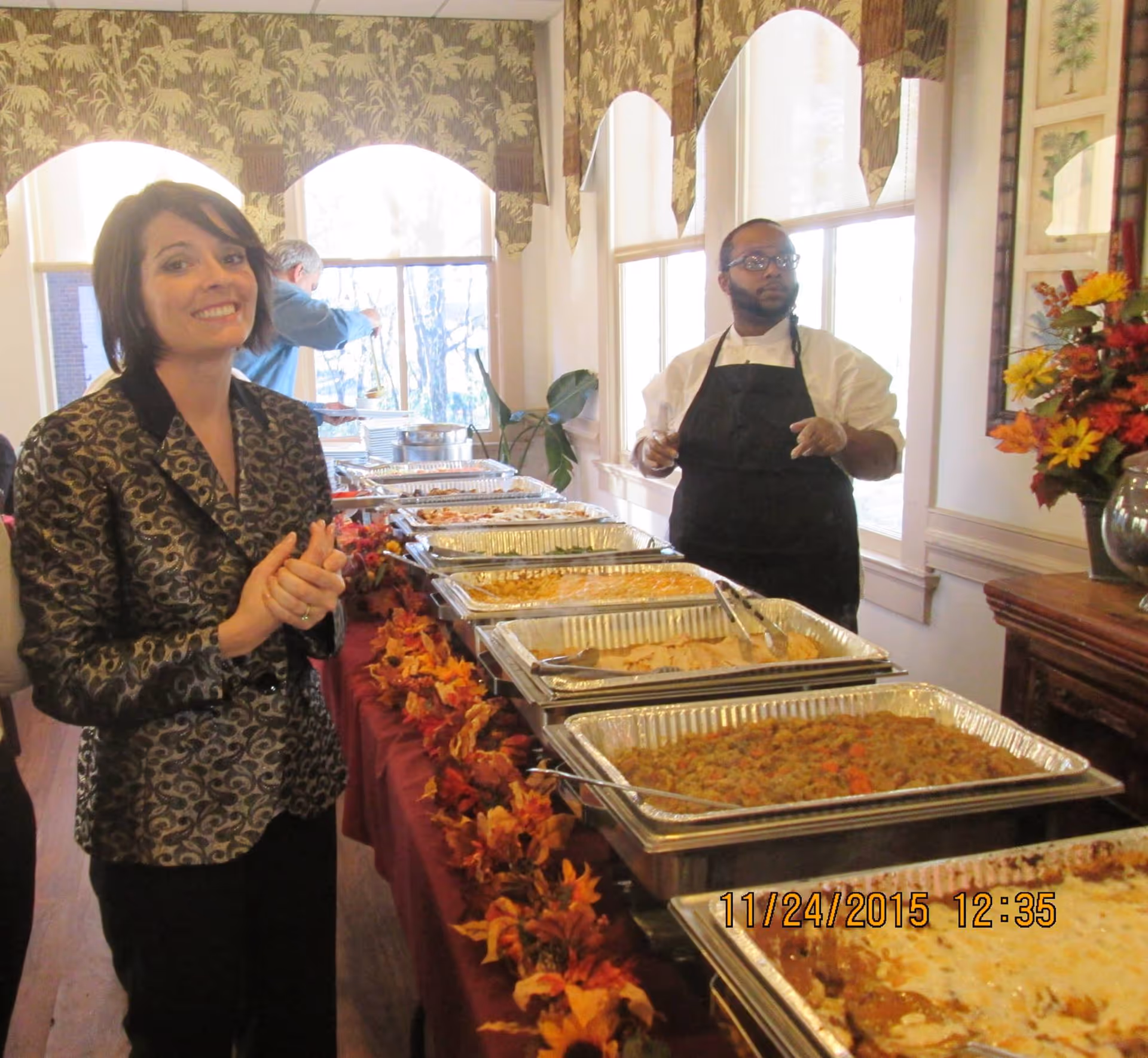 A woman smiling and standing next to a buffet table filled with trays of food in a decorated room. A man wearing a white shirt, black apron, and gloves stands behind the table near windows with patterned valances. The table is adorned with autumn-themed decorations.