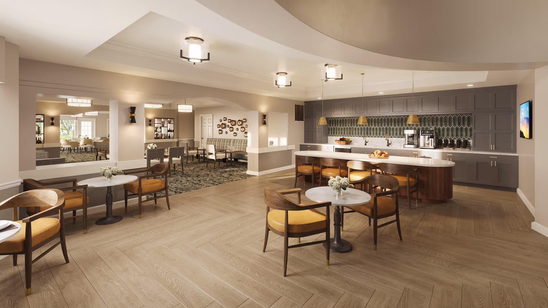 A spacious and modern common area featuring a kitchen with a large island and bar stools, several small round tables with chairs, and a seating area with a patterned carpet. The room is well-lit with ceiling lights and pendant lamps over the kitchen island. The walls are painted in neutral tones, and there is a decorative wall art piece and a flat-screen TV mounted on the wall.