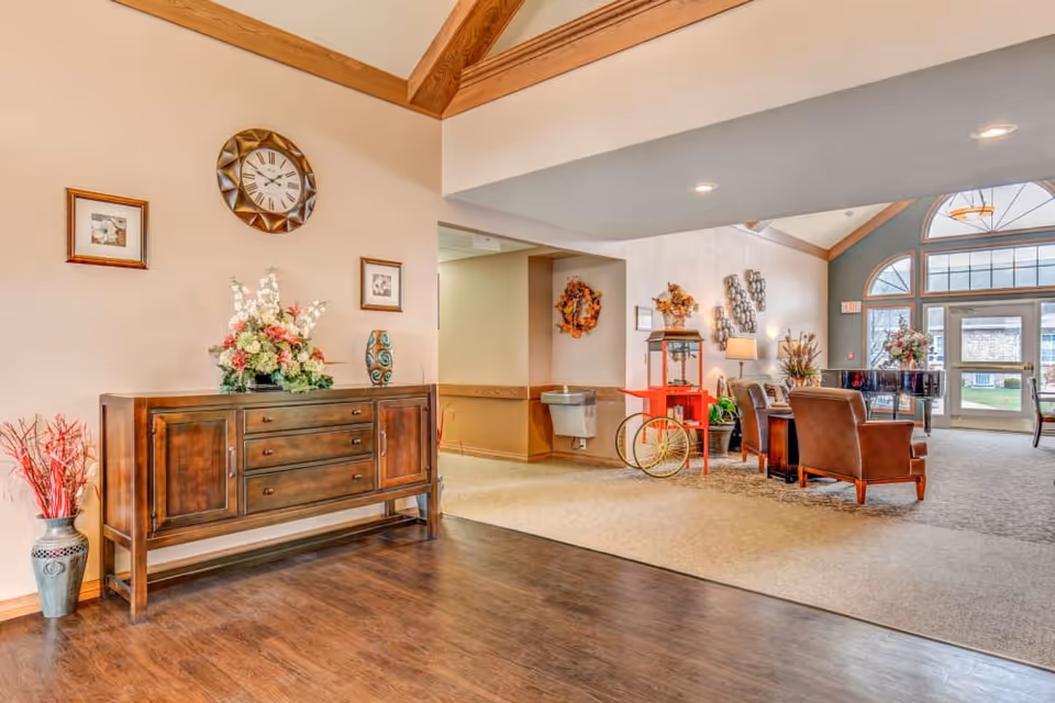Bright senior living facility lobby with a wooden sideboard and floral arrangement in the foreground and a seating area by large arched windows and entry doors.