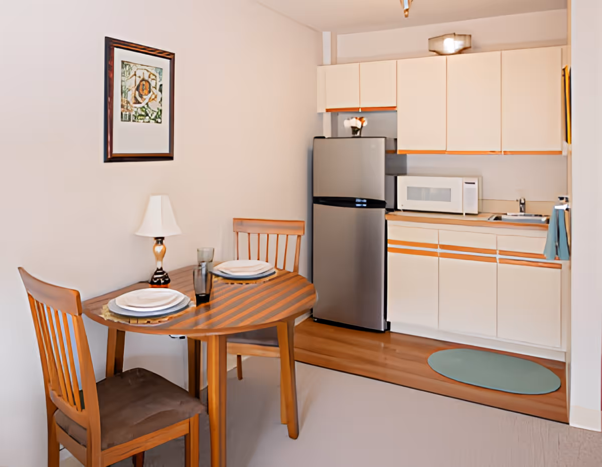 Small kitchenette and dining nook with a round wooden table set for two, stainless refrigerator, microwave, and light-colored cabinets.