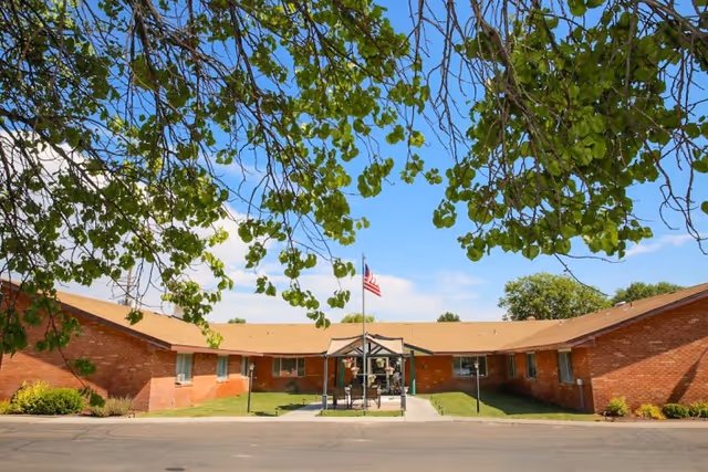 Exterior view of a single-story brick building with a tan roof, surrounded by green grass and trees with green leaves. There is a paved driveway or parking area in front, and a flagpole with an American flag in the center near a small covered seating area.
