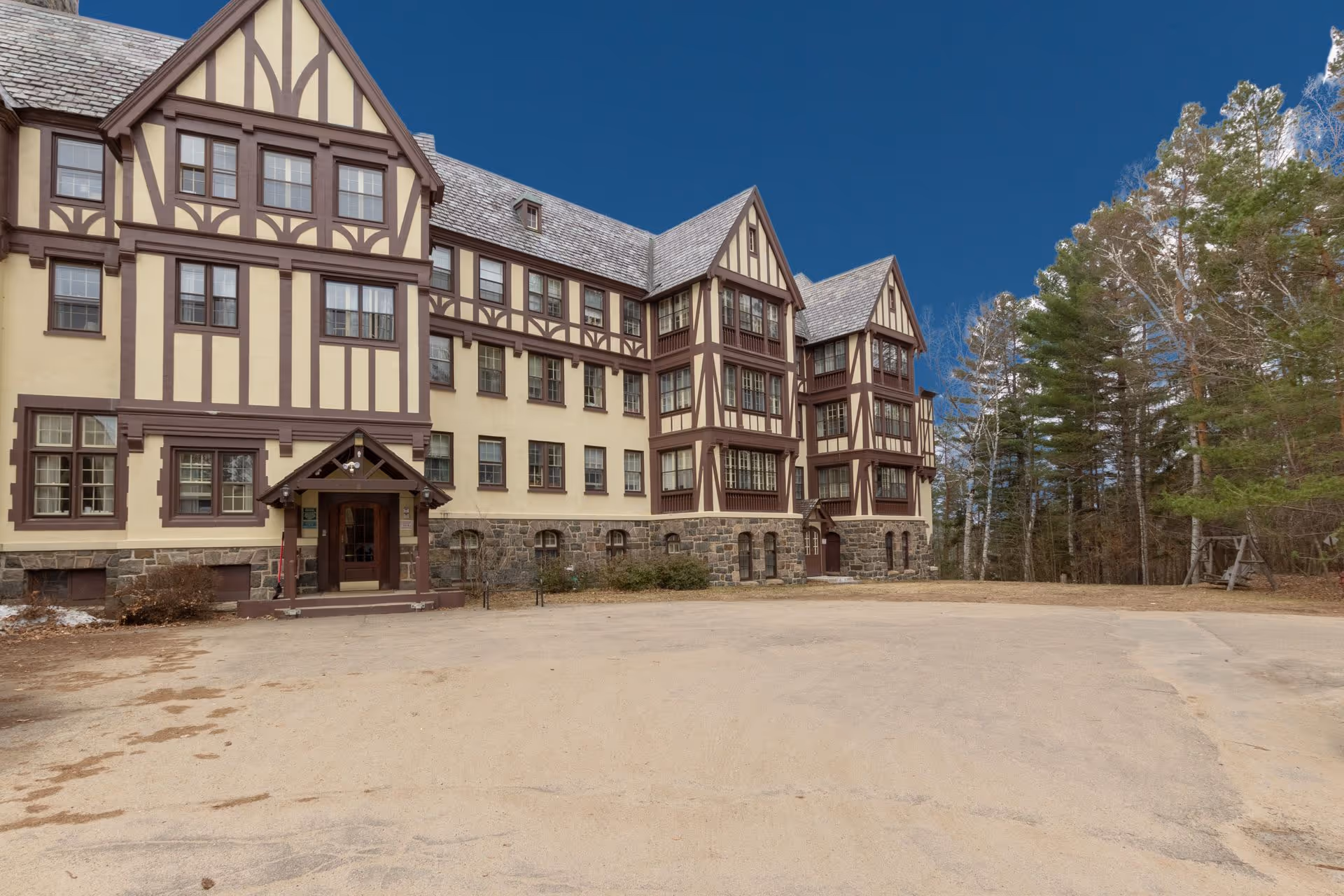 Exterior view of a large multi-story building with Tudor-style architecture, featuring beige walls with dark brown wooden beams and a stone foundation. The building is surrounded by trees and has a clear blue sky above. There is a paved area in front of the building entrance.