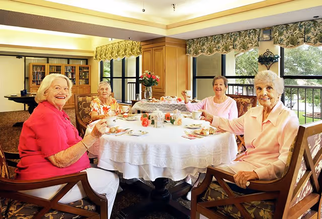 Four elderly women sitting around a round table with a white tablecloth, enjoying tea and pastries in a bright room with large windows and floral curtains. The room has warm lighting and wooden furniture, creating a cozy and inviting atmosphere.