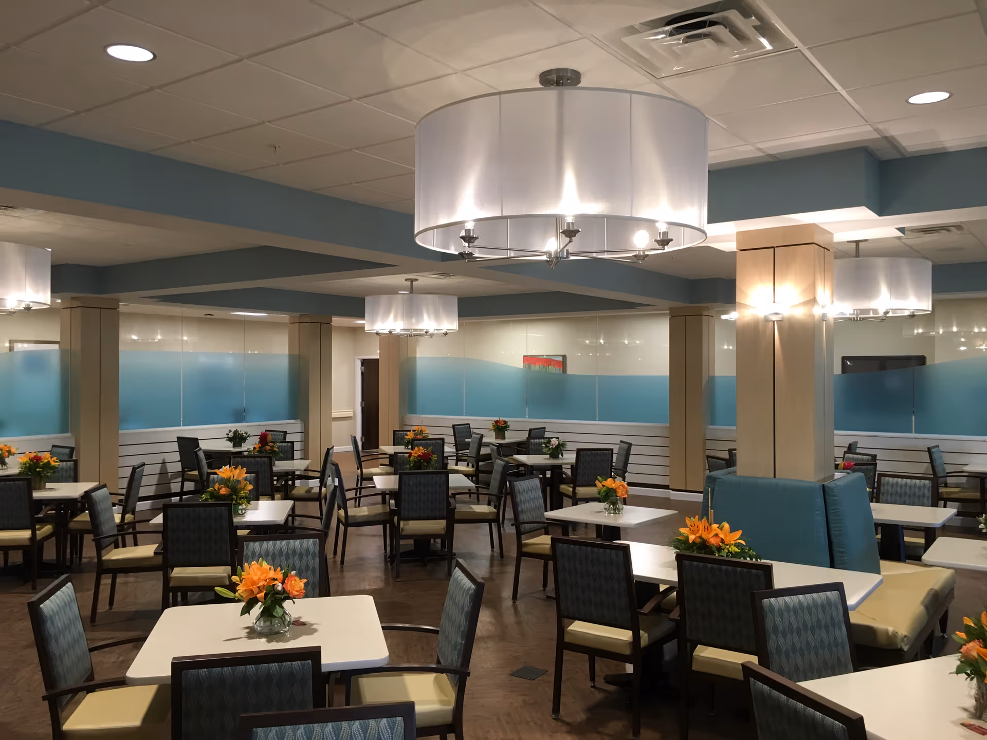 A spacious dining room with multiple square tables and chairs arranged neatly. Each table has a small vase with orange flowers. The room has a modern design with blue and beige tones, large ceiling lights, and frosted glass panels along the walls.
