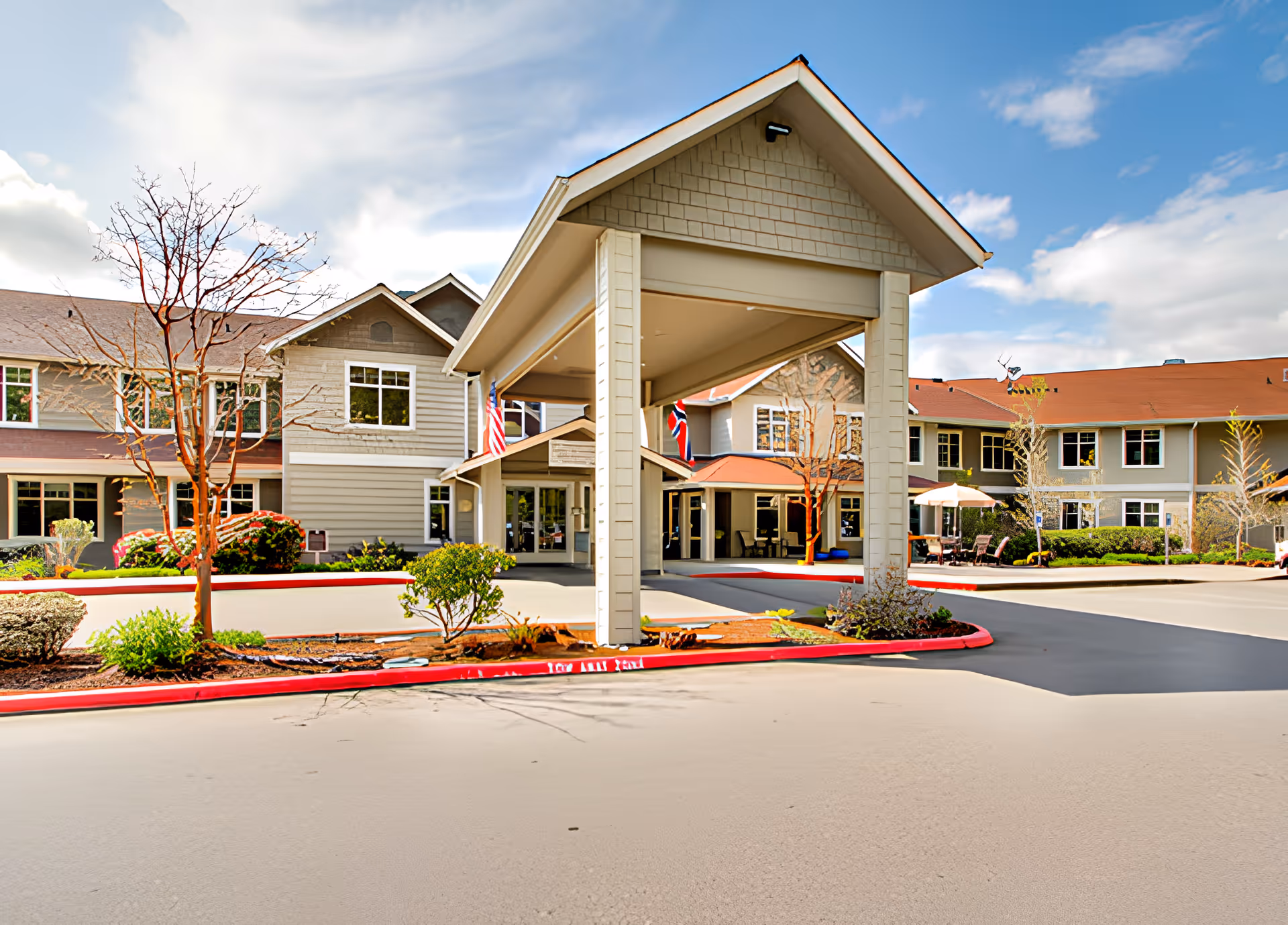 Exterior view of Brookdale Montclair Poulsbo senior living facility showing the main entrance with a covered driveway, landscaped bushes and trees, and a clear blue sky with some clouds.
