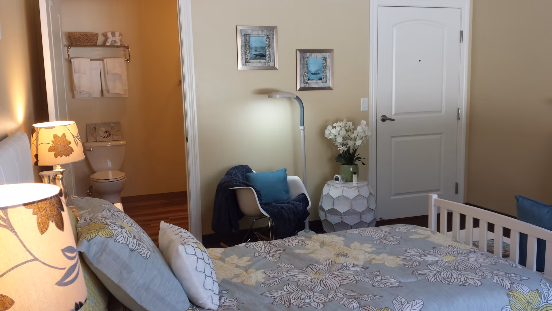 A decorated bedroom with a floral bedspread, bedside lamps, a chair and side table with flowers, and a doorway leading to a bathroom.