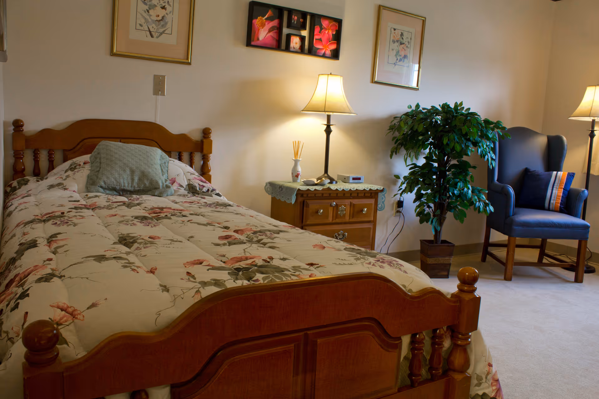 A cozy bedroom with a wooden bed frame and floral bedspread, a nightstand with a lamp and decorative items, a potted plant, and a blue upholstered armchair with a striped pillow. The walls are decorated with framed pictures and the room is softly lit by two lamps.