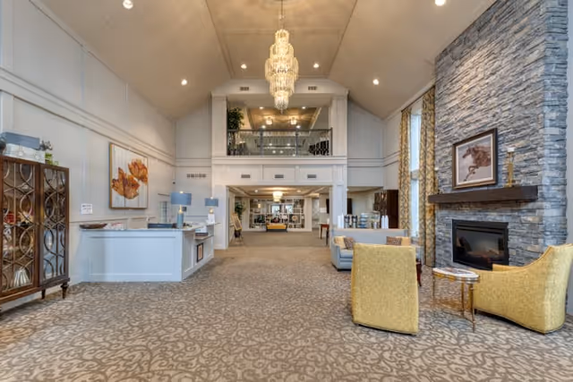 Spacious senior living facility common area with high vaulted ceilings and a large chandelier. The room features a stone fireplace with a framed picture above it, two yellow armchairs, a small round table, and a light blue sofa. There is a reception desk on the left side, decorative wooden cabinet, and large windows with patterned curtains allowing natural light to enter. The area extends into another room with additional seating and decor.